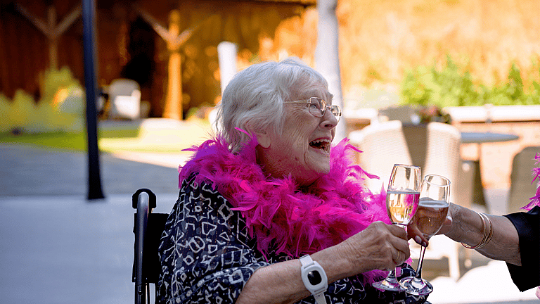 An elderly woman in a wheelchair laughs joyfully as she clinks glasses, wearing a bright pink feather boa during a lively outdoor celebration.