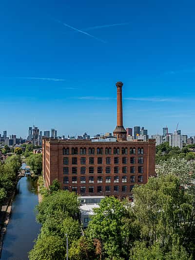 A historic red brick factory building with a tall chimney, set against the modern skyline of Manchester.