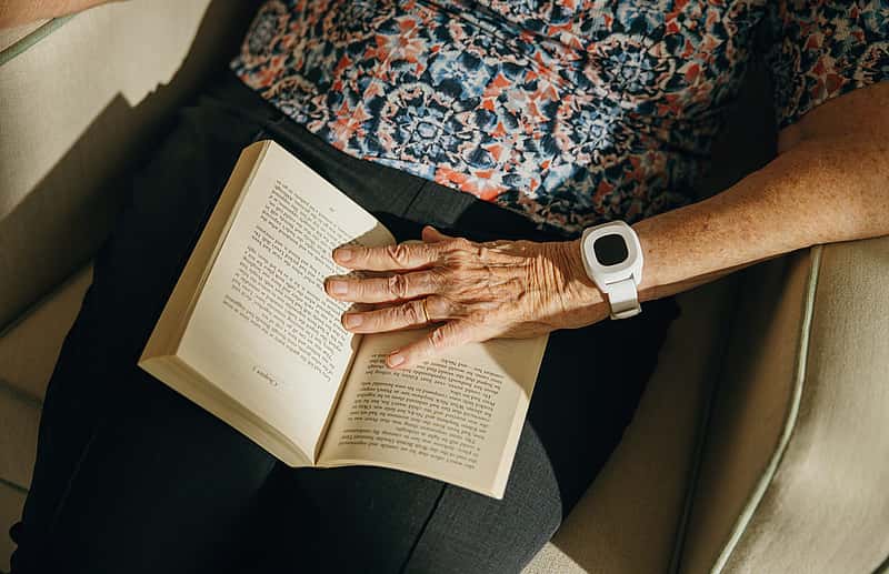 An elderly person wearing a white wrist device is sitting on a chair, resting a hand on an open book in their lap