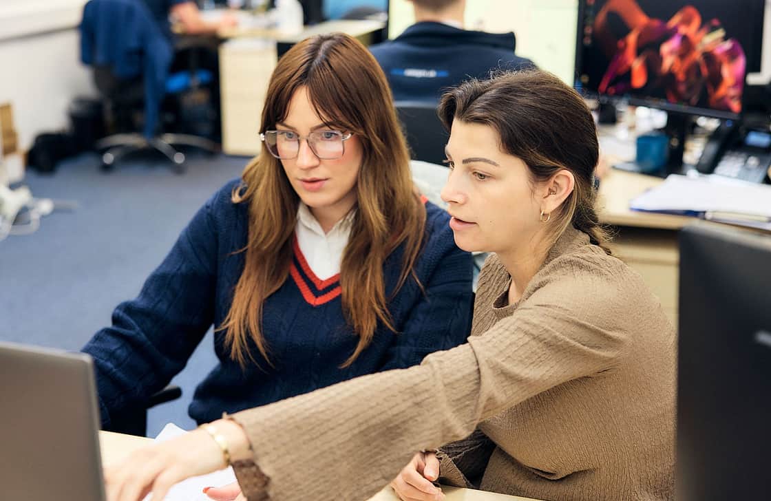 Two female team members explaining something a screen to each other sitting in an office environment.