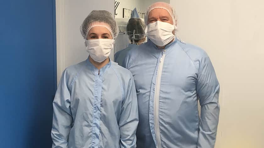 a man and a woman both in blue protective overcoats, with hair nets and face masks standing next to each other looking at the camera