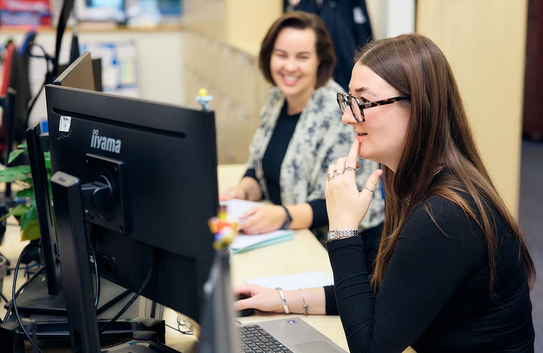Female with brown long hair wearing glasses and a black top looking at computer screen thinking with a female colleague in the background smiling