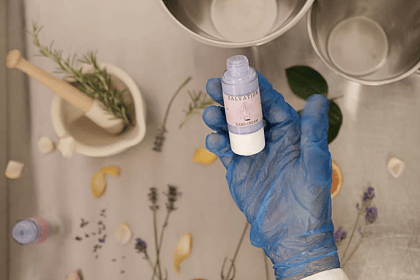 Person wearing blue gloves holding a glass cream cosmetic bottle. Above a background full of flowers and dried fruits.
