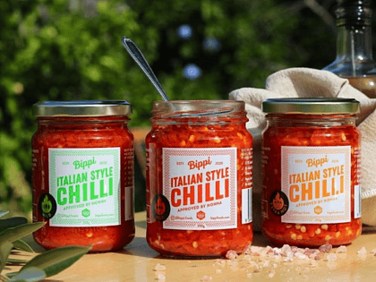 a row of three glass jars with red sauce in. The two outer jars have gold lids on, the middle jar has it's lid off and a spoon handle sticking out the top