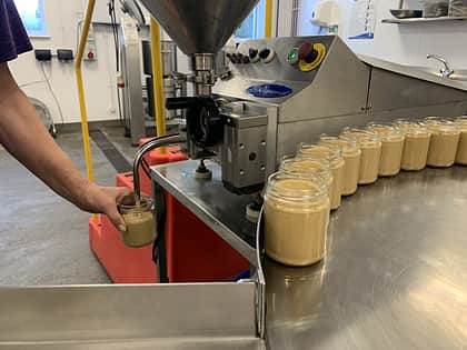a line up of glass jars with peanut butter in on a metal circular surface and a person reaching in shot holding a jar under a nozzle, which is filling the jar with the peanut butter