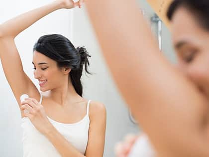 the reflection of a woman with brown hair tied up in a messy bun applying a white deodorant stick to her underarms, while smiling