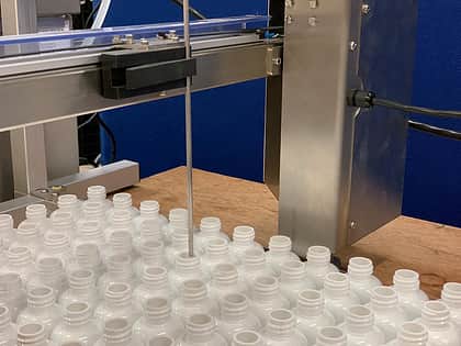 a shot of a large group of white container bottles beneath a filling machine arm, with a long thin nozzle going into the top of one of the bottles