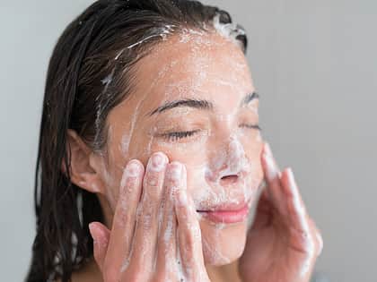 a brunette woman washing her face with exfoliator, she has her eyes closed