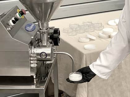 an arm reaching down holding a flat clear container, to a nozzle on a Response Benchtop filler machine, which is filling the container with a cream colour liquid