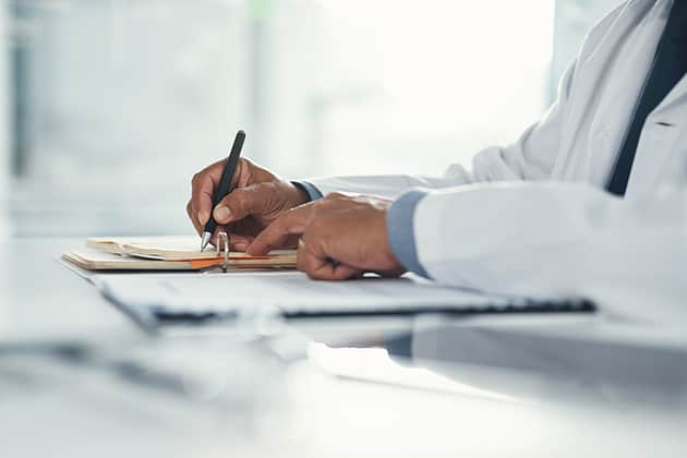 Doctor hands writing on paper or document at a desk in the hospital. Healthcare professional drafting a medical insurance letter, legal paperwork or form. A GP filing a document in a clinic office
