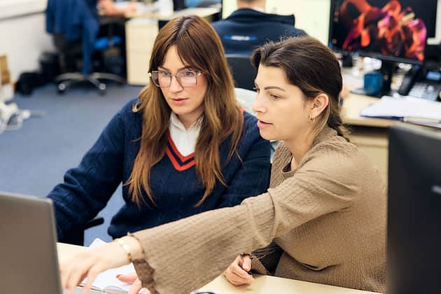 Two female team members explaining something a screen to each other sitting in an office environment.