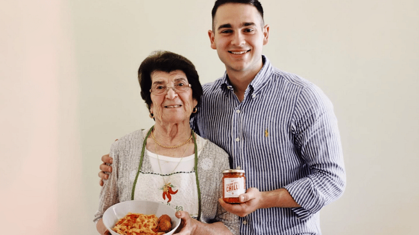 a young man with brown hair and wearing a blue and white stripe shirt holding a glass jar filled with red sauce, has his arm around an older lady with brown hair, holding a white bowl with a pasta and meatballs food in. She also has brown hair and wearing glasses.