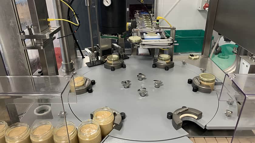 factory machinery with a line up of jars filled with a peanut butter waiting to enter onto a circular floor to then have the lids applied