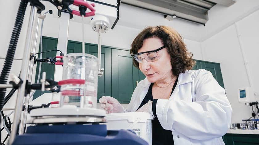 a brunette haired woman in a lab coat, in a laboratory setting leaning over a desk working, with a clear glass beaker in the foreground