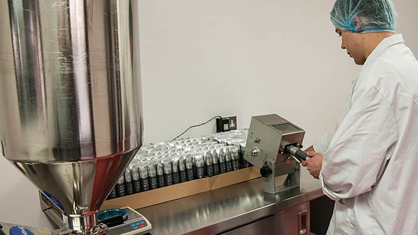 a large silver metal funnel on the left side with a collection of small container bottles behind the machine. A man in a white lab coat with a blue hair net is standing working with a machine on the right side