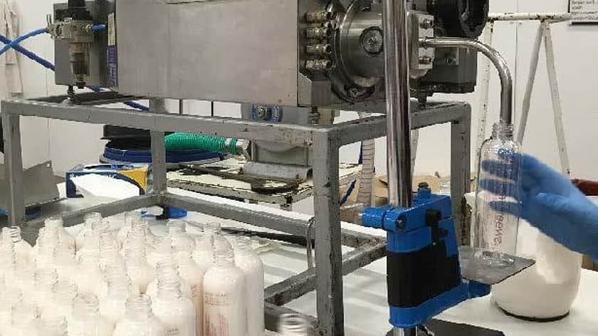 someone wearing blue protective blue gloves in the process of filling a collection of clear bottles with a cream from a filling machine in a factory setting