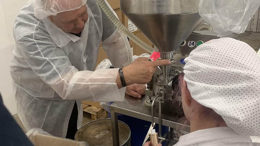 a woman in a white lab coat and hair cover is kneeling with her back to the camera, while a man also in a white lab coat and hair cover leans in to use a Response Benchtop filler