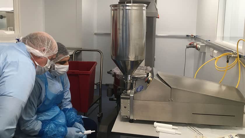 two people in blue protective overcoats, with hair nets and face masks bend down to look at a Response Benchtop Filler machine