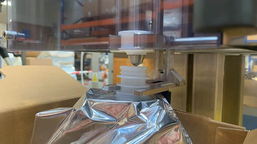 a close up of a silver bag container underneath a filling nozzle, the top of the bag opening being held in place with metal holder