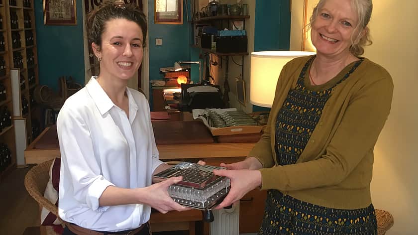 two women smile at the camera, one wearing a white shirt and the other a green dress with green cardigan, who is handing over a ProFiller 1100