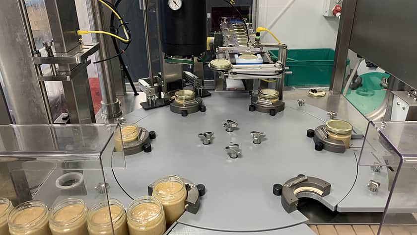 factory machinery with a line up of jars filled with a peanut butter waiting to enter onto a circular floor to then have the lids applied