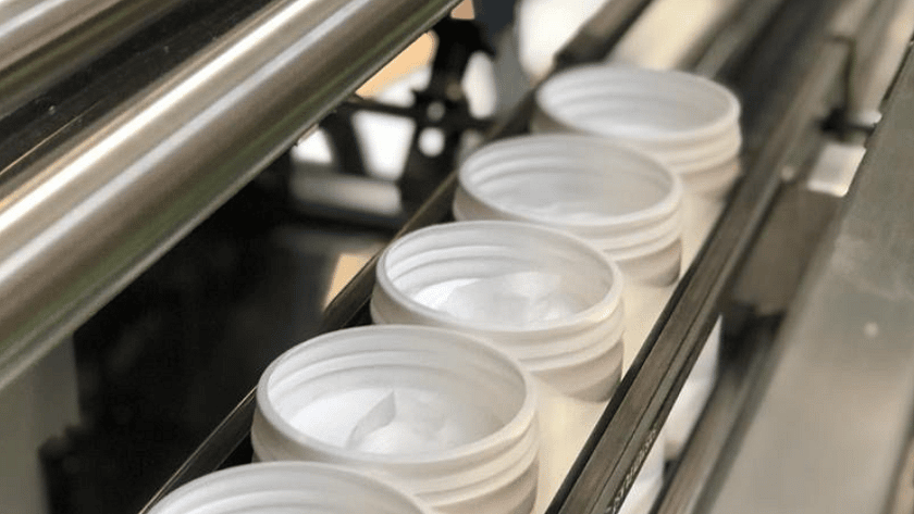A close up of line up of white tubs in a filling machine, all filled with a white cream