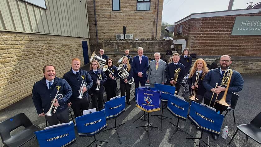 A group of 13 people stand facing the camera, 11 of whom are holding brass band instruments. Chris Wilson and Stephen Holroyd stand in the centre of the group. There are a number of music stands in front of the group, with blue and gold banners hanging off the front, with the word 'Hepworth' embroidered on in gold.