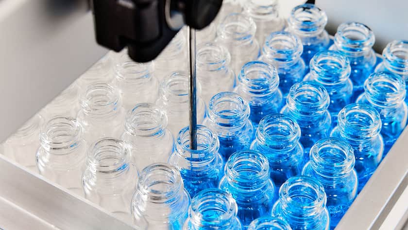 Liquid dispensing nozzle filling glass vials with blue solution on a stainless steel tray, highlighting precision dosing, automated filling, and pharmaceutical or cosmetic production processes.