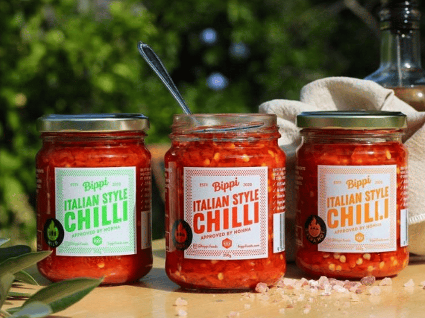 a row of three glass jars with red sauce in. The two outer jars have gold lids on, the middle jar has it's lid off and a spoon handle sticking out the top