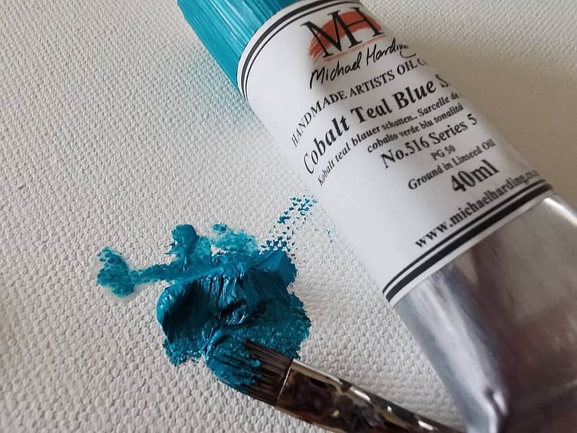 a turquoise paint tube lying on a canvas, with a paint splatter next to it and a paint brush lying in the splatter