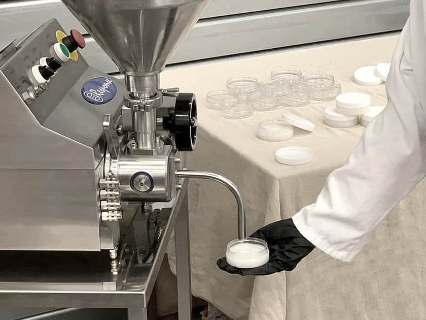 an arm reaching down holding a flat clear container, to a nozzle on a Response Benchtop filler machine, which is filling the container with a cream colour liquid