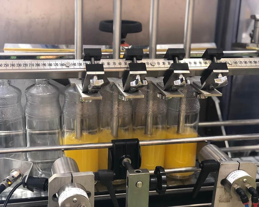 A 4 head filling machine in the middle of filling orange juice in large see through containers from the bottom up.