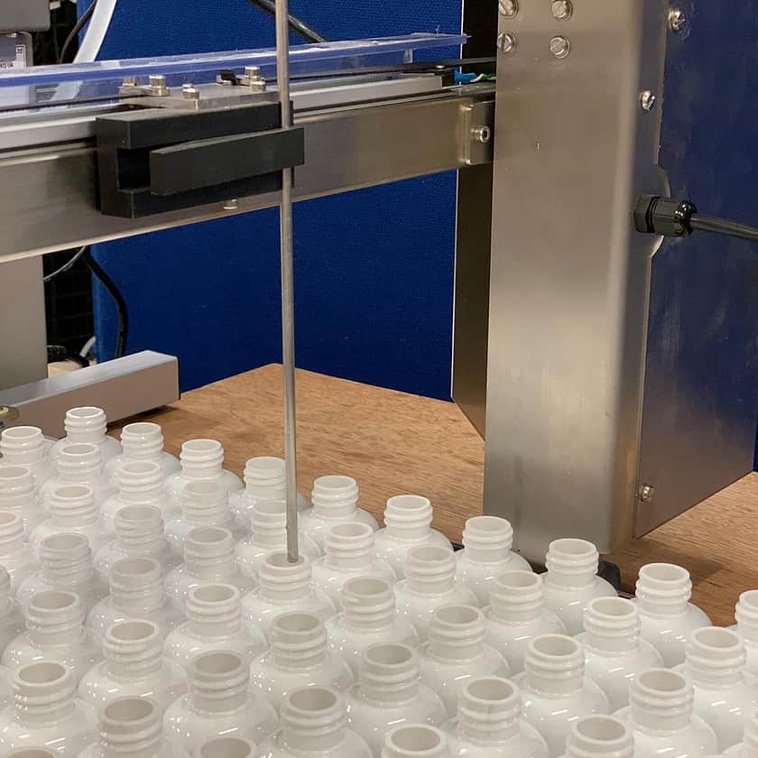 Close-up of automated bottle filling line with stainless steel dispensing nozzle positioned above rows of empty white plastic bottles, featuring precision alignment system and industrial support frame for pharmaceutical, cosmetic or chemical liquid packaging.