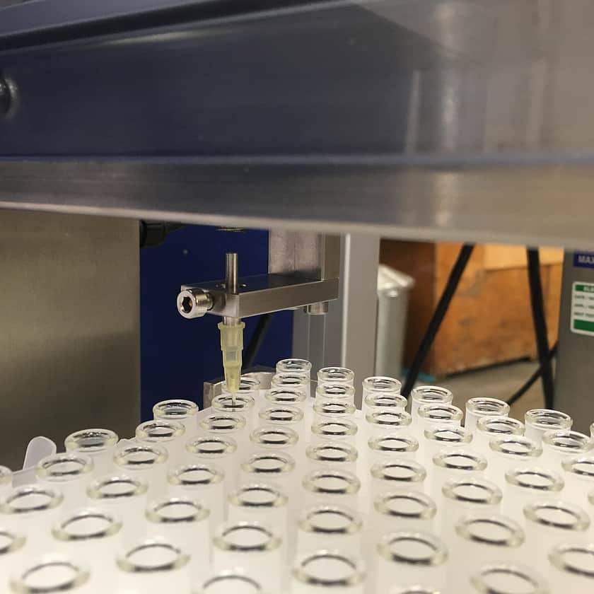 Close-up of automated liquid filling nozzle dispensing fluid into glass vials arranged in a tray, featuring precision dosing needle and stainless steel machinery for accurate pharmaceutical and laboratory packaging applications.