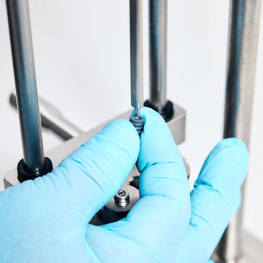 Close-up of laboratory testing equipment with gloved hand holding a small component under a precision instrument for quality control analysis