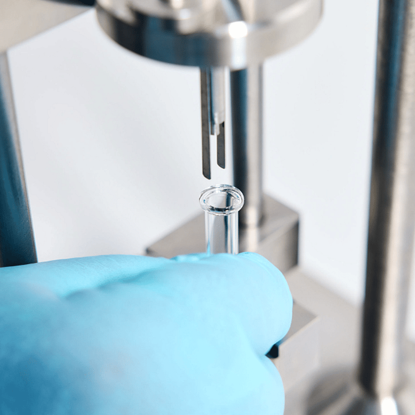 Close-up of laboratory testing equipment with gloved hand holding a glass vial under a precision instrument for quality control analysis