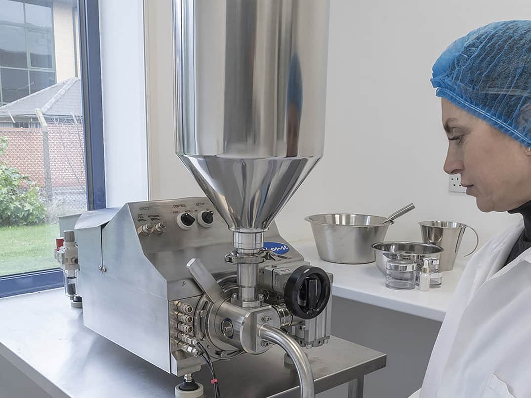 a woman with a blue hair net and white lab coat stands in front of a Response Benchtop Filler, filling up a small black container with a cream substance