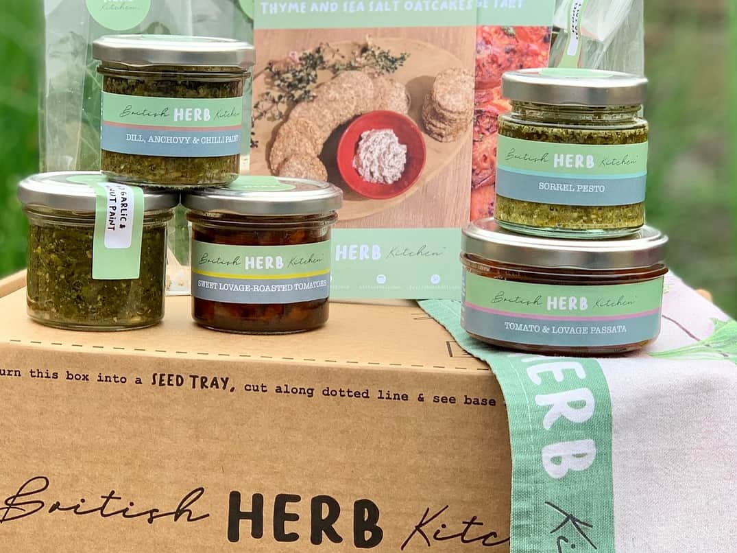 a cardboard box with the British Herb Kitchen logo printed on it, with a selection of their products sitting on top with glass jars and branded labels on them