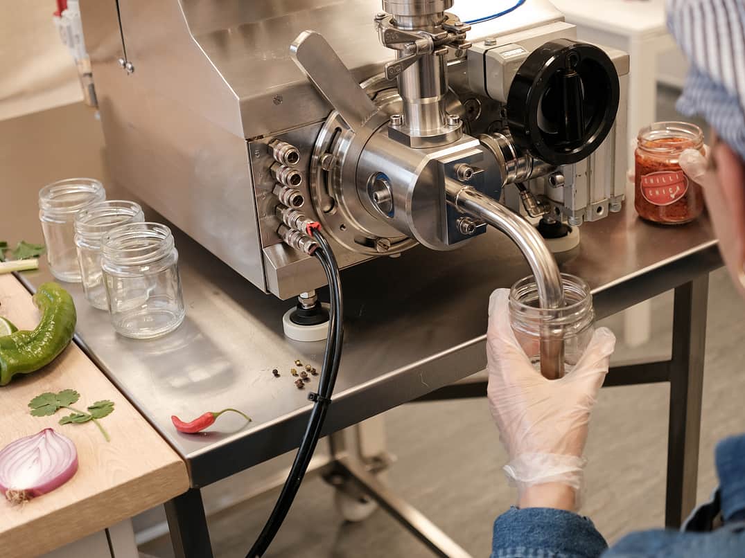 The Adelphi Bench Top Response Filler is in use and filling small pots of chilli jam. Around the bench top filler there are empty jars and chillis placed around. The filler is wearing a bakers hat.