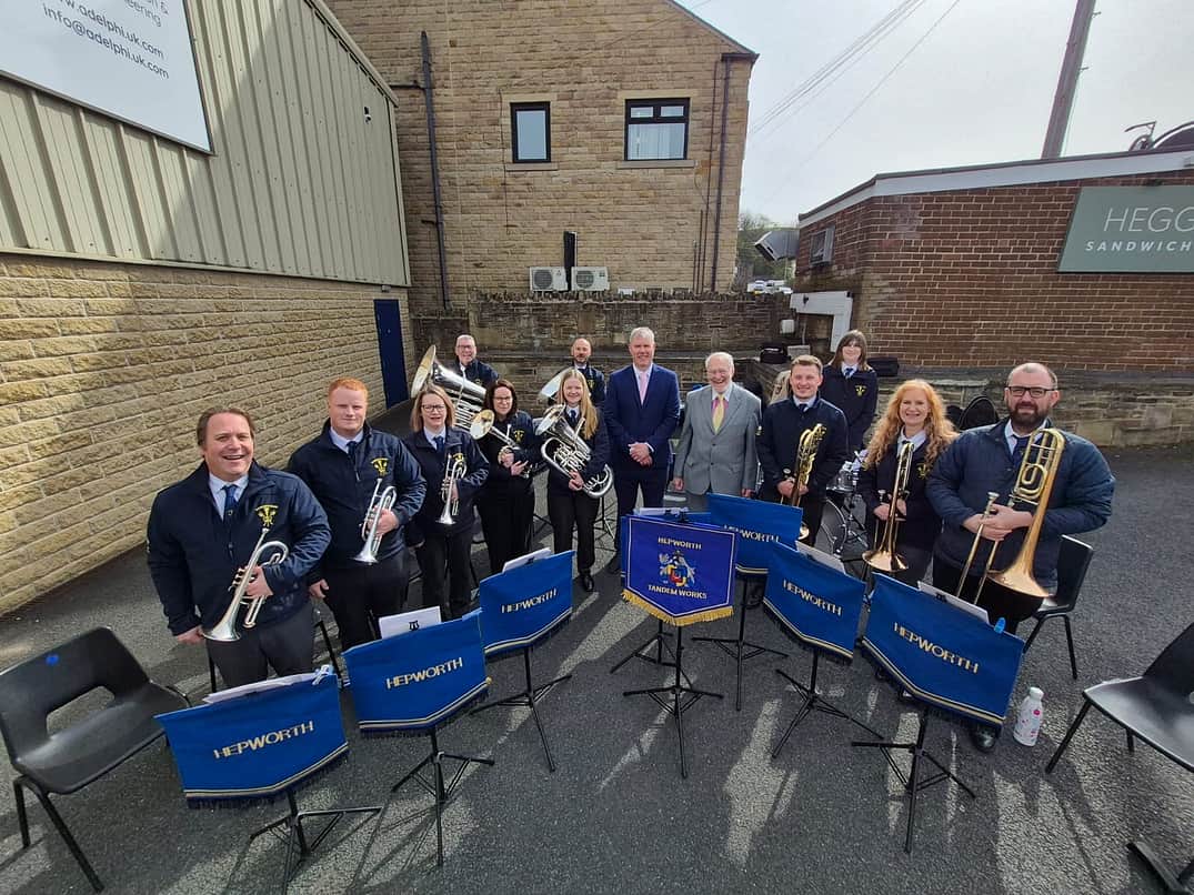 A group of 13 people stand facing the camera, 11 of whom are holding brass band instruments. Chris Wilson and Stephen Holroyd stand in the centre of the group. There are a number of music stands in front of the group, with blue and gold banners hanging off the front, with the word 'Hepworth' embroidered on in gold.