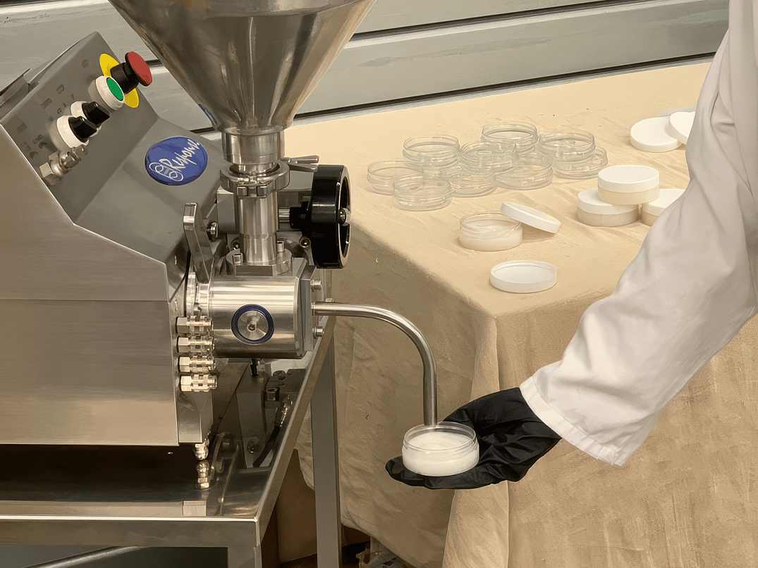 A person wearing a lab coat fills up a small container with cosmetic cream, using a Respone Benchtop Filler