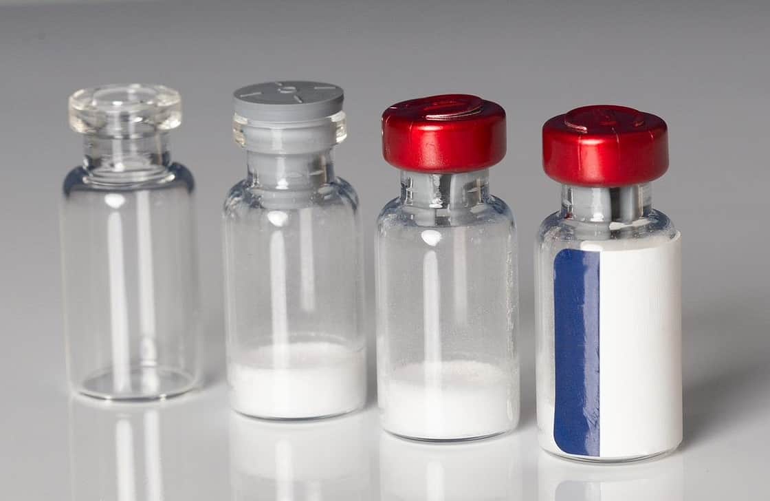 Four vials: one empty, one containing lyophilised powder with a stopper, one containing powder, with a red crimp seal and one sealed and labelled.
