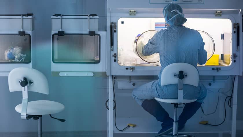 Ein Laborarbeiter sitzt auf einem Stuhl und hat seine Arme in einem aseptischen Isolator.