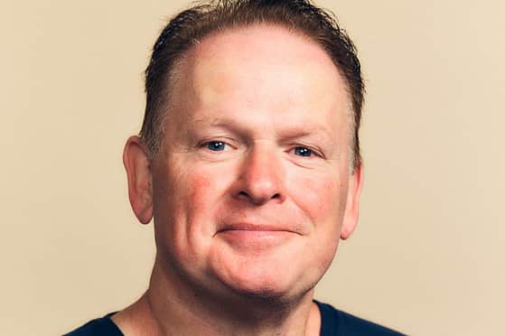 A smiling man with brown hair, wearing a navy blue t-shirt with an Adelphi logo on the chest.