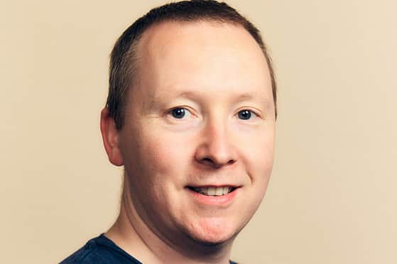 A smiling man with short hair wearing a navy blue T-shirt with an Adelphi logo on the chest.