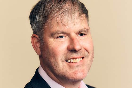 A smiling man wearing a light pink shirt and dark suit jacket.