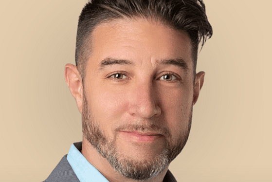 A headshot of a man with brown hair wearing a light blue shirt and a light gray blazer.