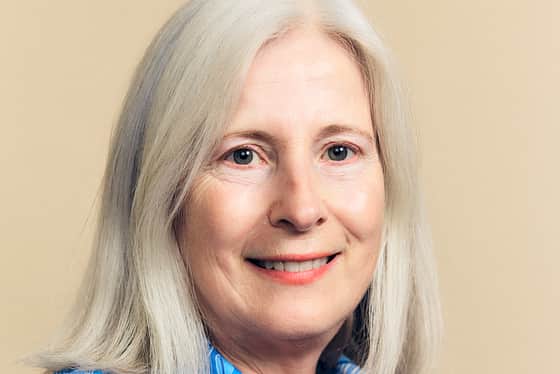 A side on profile of a female wearing a floral blue blouse. The female has a white long bob. She is smiling with her teeth and standing in front of a peachy coloured background