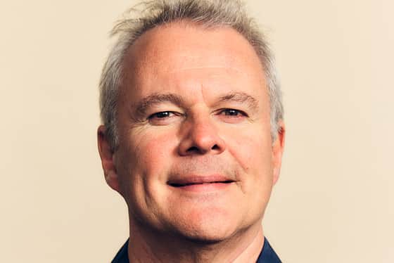 A front on profile of a man wearing a blue lightly patterned shirt. The man has short grey hair that has been swept upwards. He is smiling and standing in front of a peachy coloured background