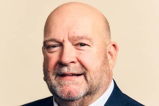 A side on profile of a man wearing a full suit with a blue tie, white shirt and a blue jacket. The man is bald and he is smiling, standing in front of a peachy coloured background